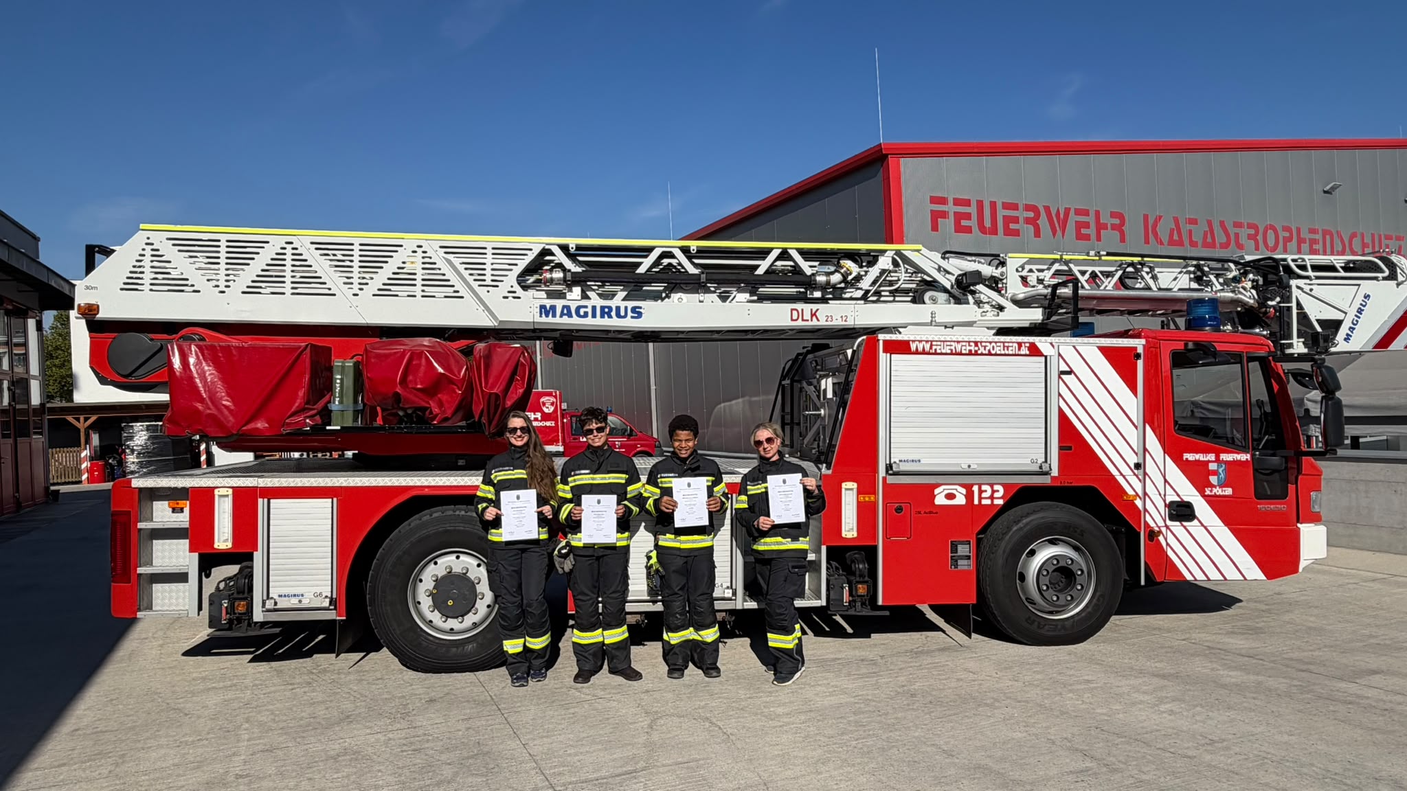 Vier junge Feuerwehrleute mit Urkunden vor einem Magirus-Drehleiterwagen der Feuerwehr St. Pölten