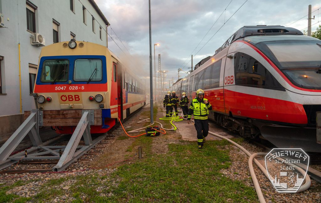 Feuerwehrleute im Einsatz bei zwei ÖBB-Zügen in St. Pölten mit Schläuchen und Rauch.